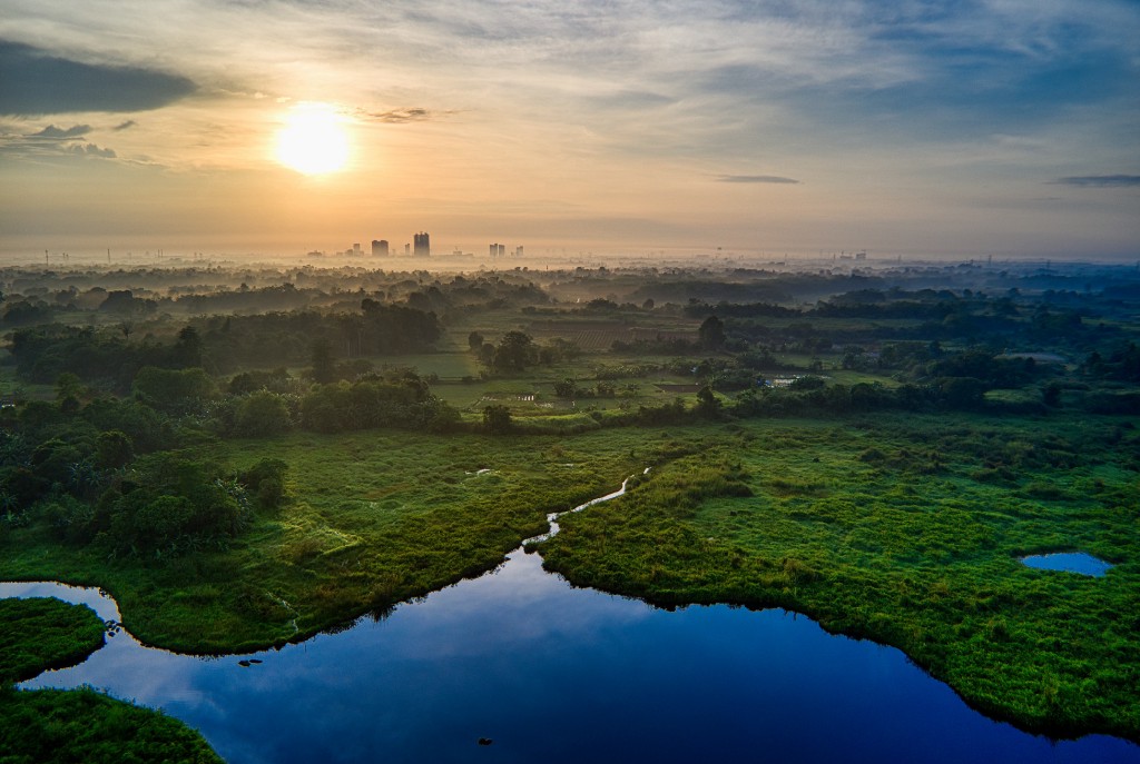 Landscape during dusk — lake in the foreground, followed by green landscape & city in the far background, sun shining bright.