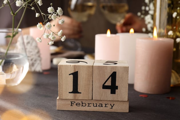 wooden blocks that read February 14 on a dinner table with candles, flowers, and a couple toasting in the background