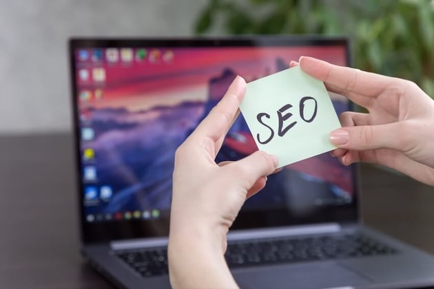 Person holding a Post-it note that says SEO in front of a MacBook