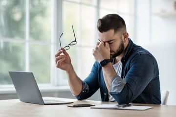 Frustrated young man in shirt, with laptop at remote job, takes off glasses, rubs eyes