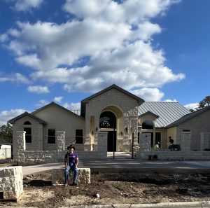 Patrick in front of his newly built home sitting on a concrete slab in the front yard. The home is mostly complete but there is still some landscaping work in the front yard to be completed.