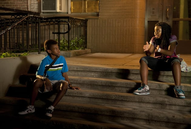 two young Black people sit on stems in front of an apartment building