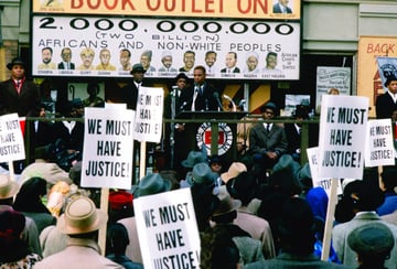 several Black men stand on a stage with a podium in front of a large crowd with signs saying 'we must have justice'